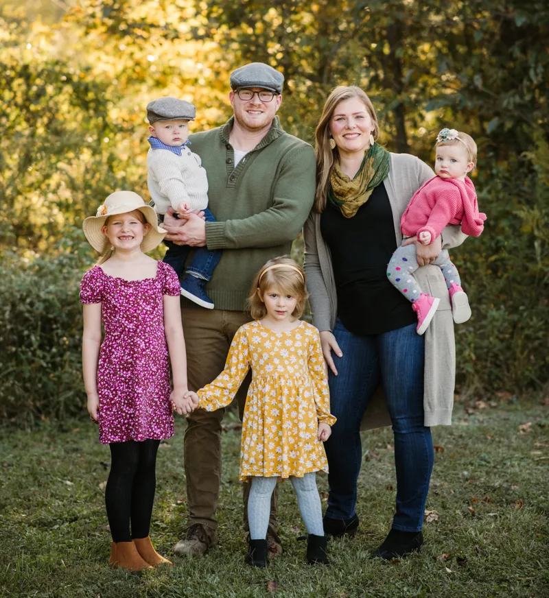 Christina with her husband and their four children, standing together outdoors among autumn trees.